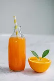 A glass bottle of orange juice with a paper straw standing on marble, beside a fresh orange cut in half with two leaves.