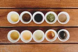 Ten small white bowls in two rows on a wooden table, holding syrups and jams in amber, green, caramel and dark molasses tones.