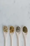 Four wooden spoons in a row on white marble, each holding a different dried botanical: chamomile, lavender, mint and crushed leaf.