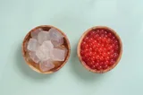 Two wooden bowls seen from above on a mint green surface: translucent jelly cubes in syrup on the left, red popping boba on the right.